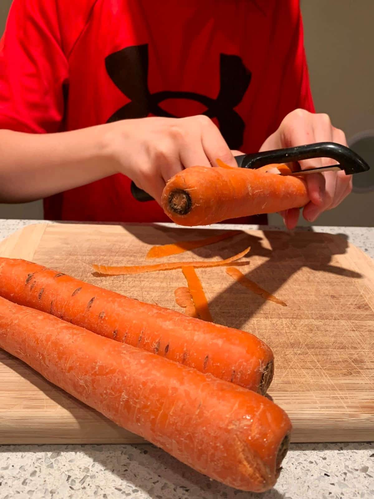 child peeling carrots with vegetable peeler over wooden cutting board