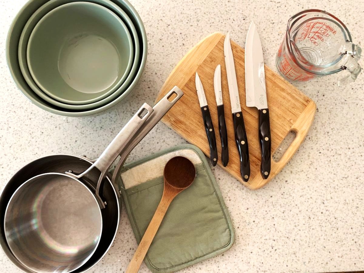 Essential cooking gear for kids including glass mixing bowls, stainless steel pots and pans, hot pad, wooden spoon, wooden cutting board and knives